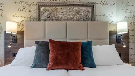 A neatly made bed with a white quilt and three velvet pillows against a cushioned headboard, two wall-mounted lamps, and a framed map above.
