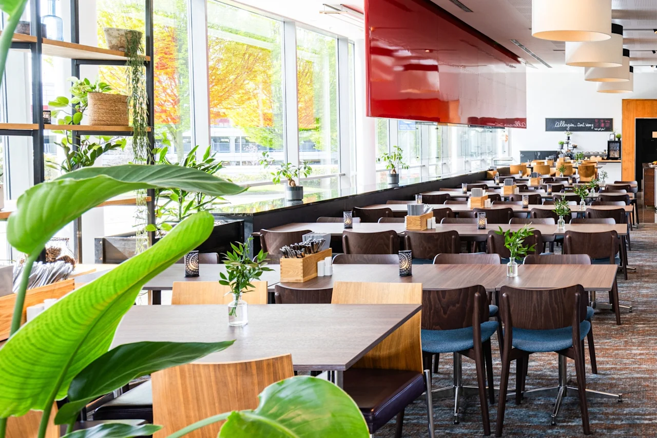 Bord og stoler fyller en romslig, lys restaurant dekorert med grønne planter og store vinduer. En tavle viser teksten: "Breakfast served from 7am - 10am."
