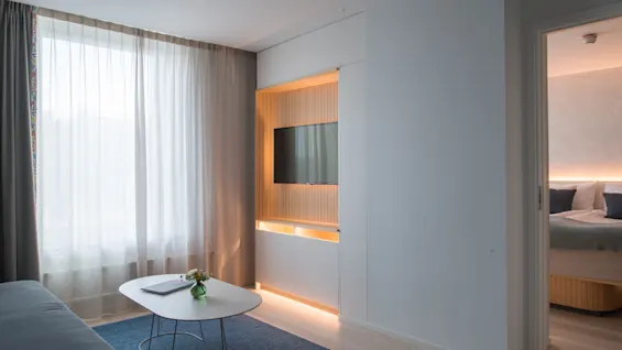 A modern living room features a white coffee table with a green vase and book, positioned on a blue rug. Nearby, a wall-mounted TV sits in a lighted alcove beside a window.