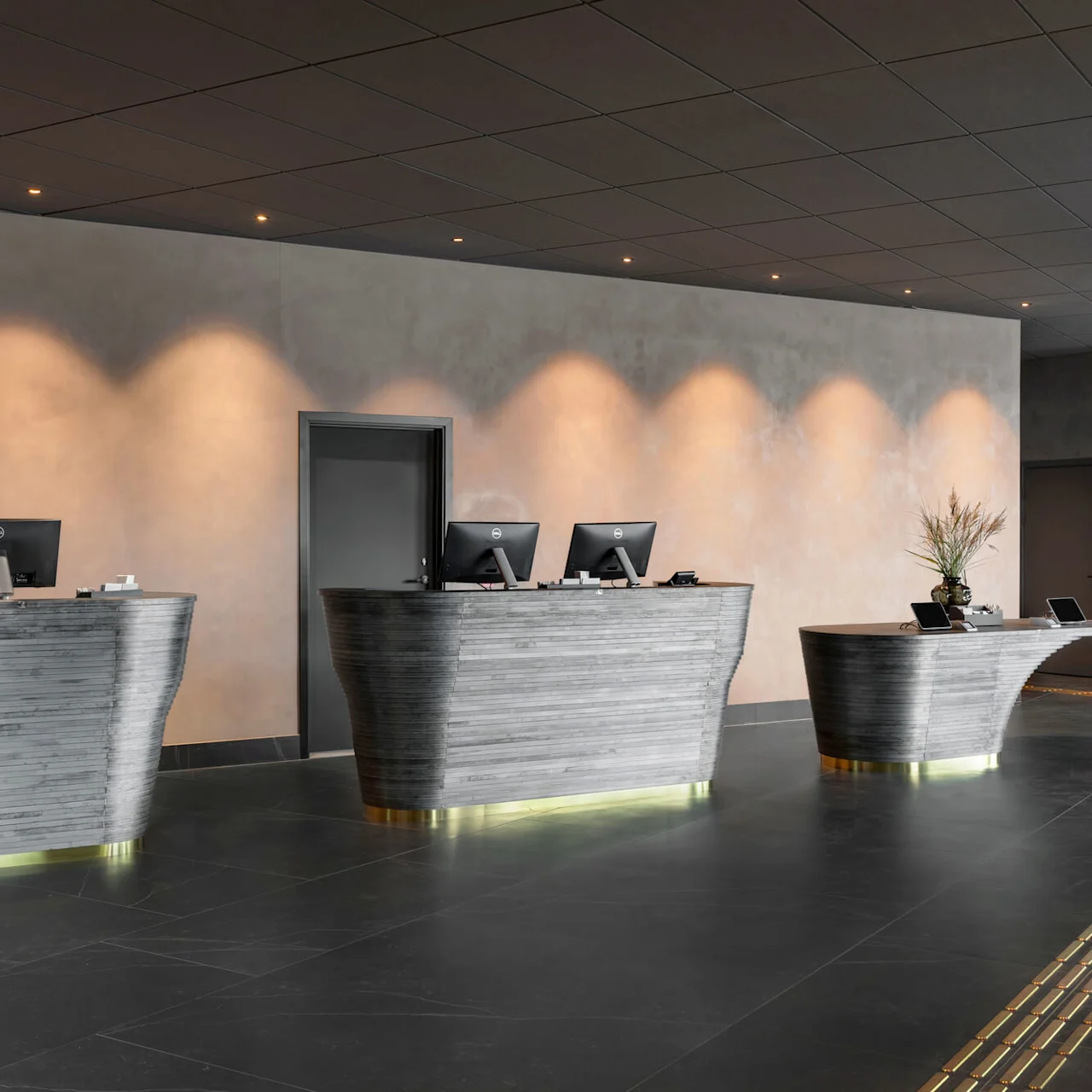 Tre moderna receptionsdiskar med datorer står obemannade i en snygg, minimalistisk lobby, upplysta av taklampor och flankerade av en hylla fylld med flaskor.