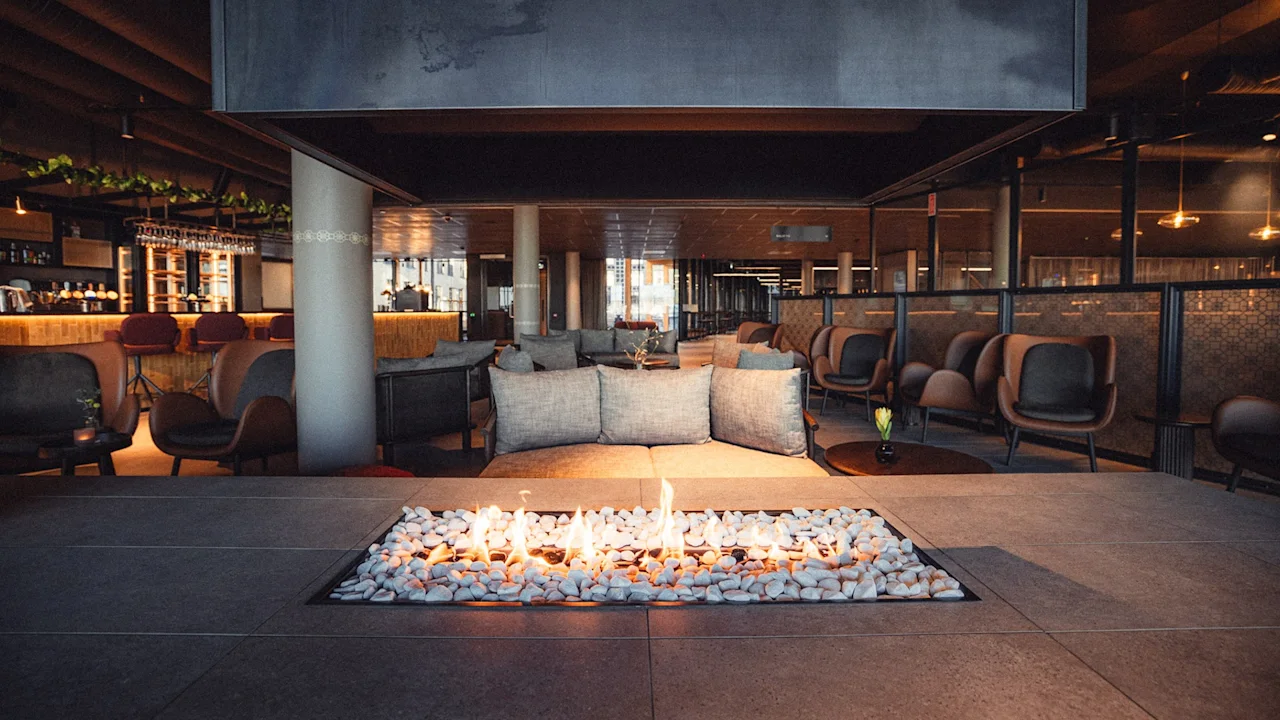 En moderne lounge har en rektangulær ildsted i midten med flammer over sten, omgitt av stoler og sofaer. Miljøet er en stilren innendørs arena med varm belysning.
