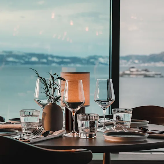 Et bord er elegant dekket med glass, tallerkener og en liten plante, plassert ved et stort vindu som viser en rolig utsikt over havet og fjerne åser.