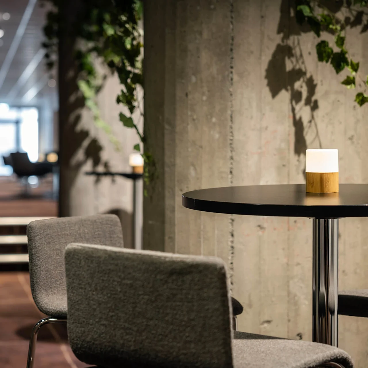 Et rundt bord med en lille lampe på toppen er omgivet af grå stole, i et moderne, blødt belyst interiør med betonvægge dekoreret med grønt.