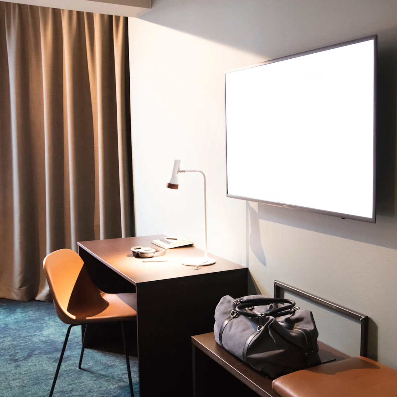 A wall-mounted TV emits light in a hotel room. A desk with a lamp, chair, and papers is nearby. A duffel bag rests on a bench under the TV.