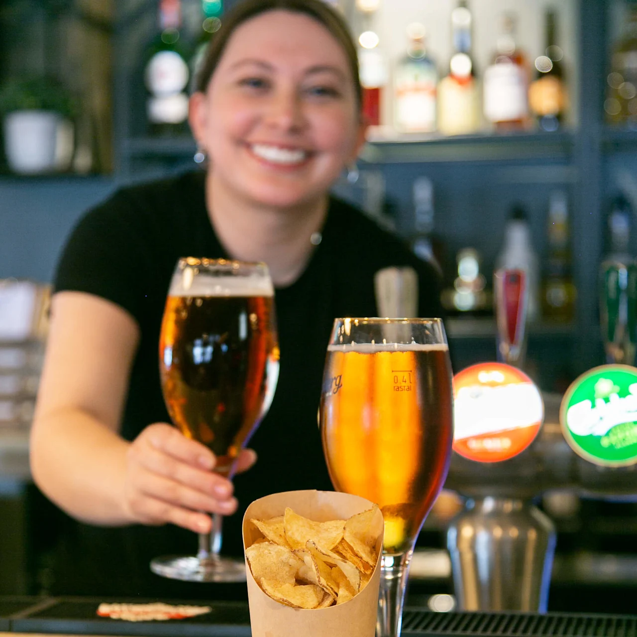 To glas øl står på en bardisk, ledsaget af en papirkop med chips. En smilende bartender i baggrunden holder et glas. Barsætning med flasker synlige.