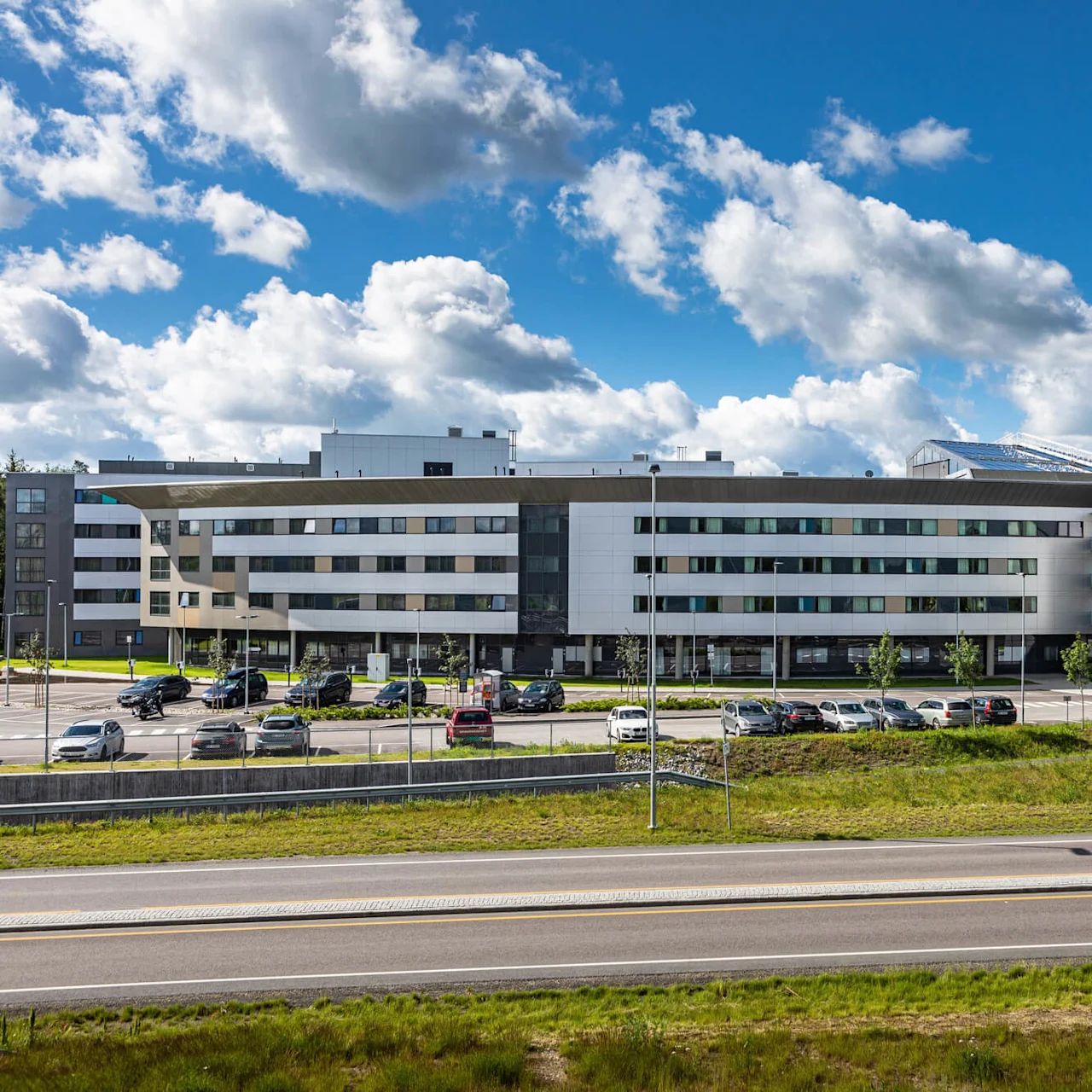Quality Airport Hotel Gardermoen stands with modern architecture amid parked cars, under a bright blue sky with clouds, surrounded by trees and a grassy area alongside a road.