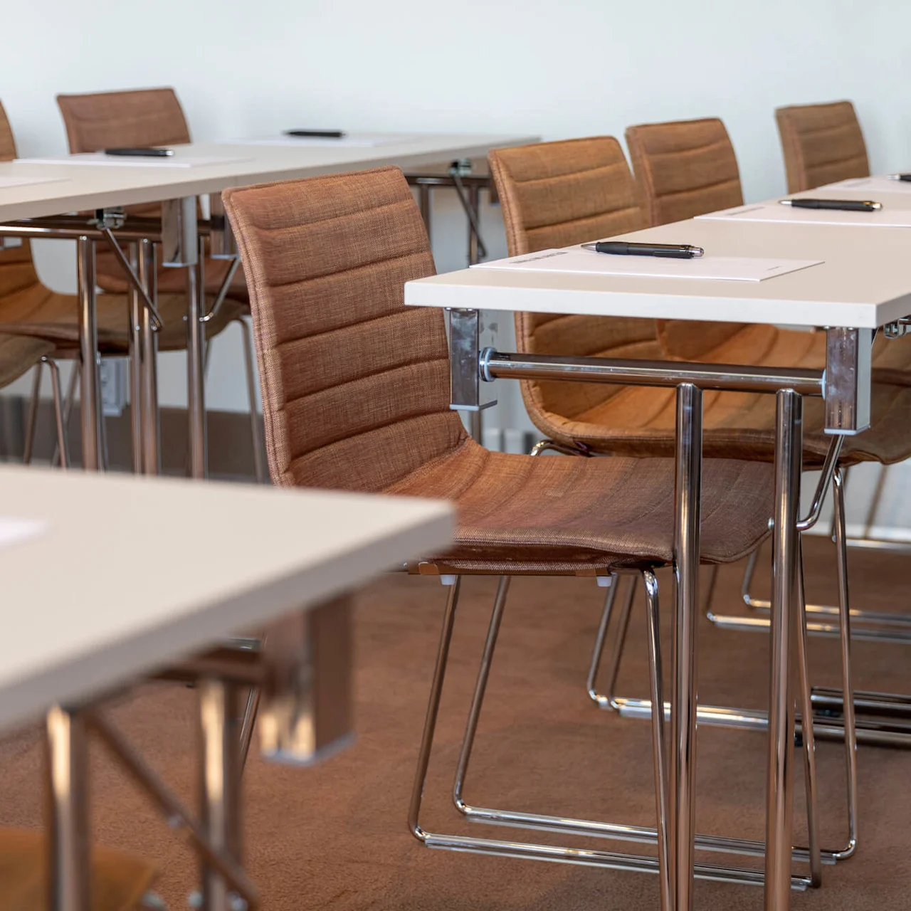 Desks with paper and pens are arranged in neat rows, paired with brown chairs. They are set in a carpeted classroom or conference room.