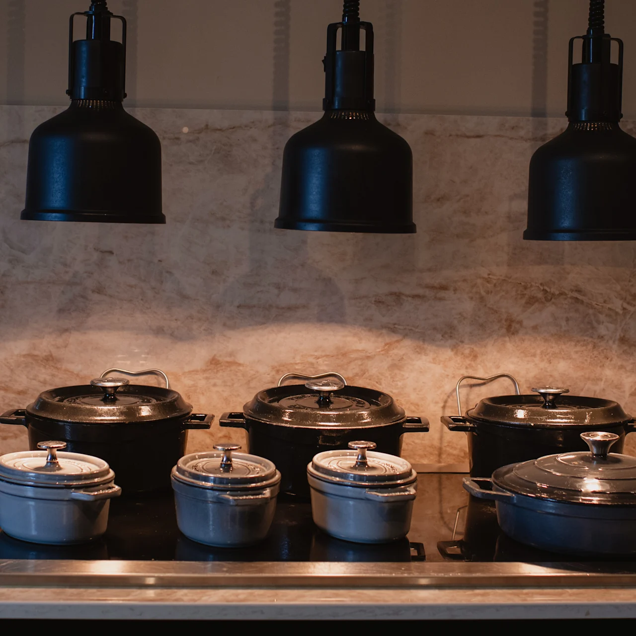 Black and white pots with lids rest on a modern stovetop. Three overhead lights illuminate the kitchen area, which features a marble backsplash.