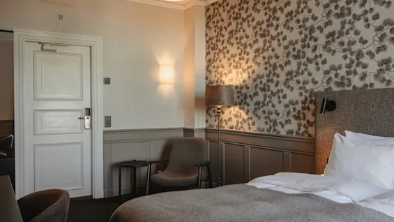 A cozy hotel room with a bed, chair, and lamps, featuring patterned wallpaper and a warm ambiance. Home Hotel Post.