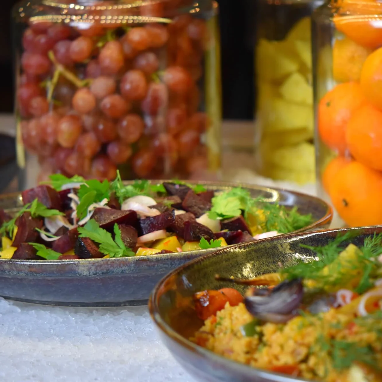Two bowls of colorful salads on a bed of ice, with jars of preserved fruits in the background.