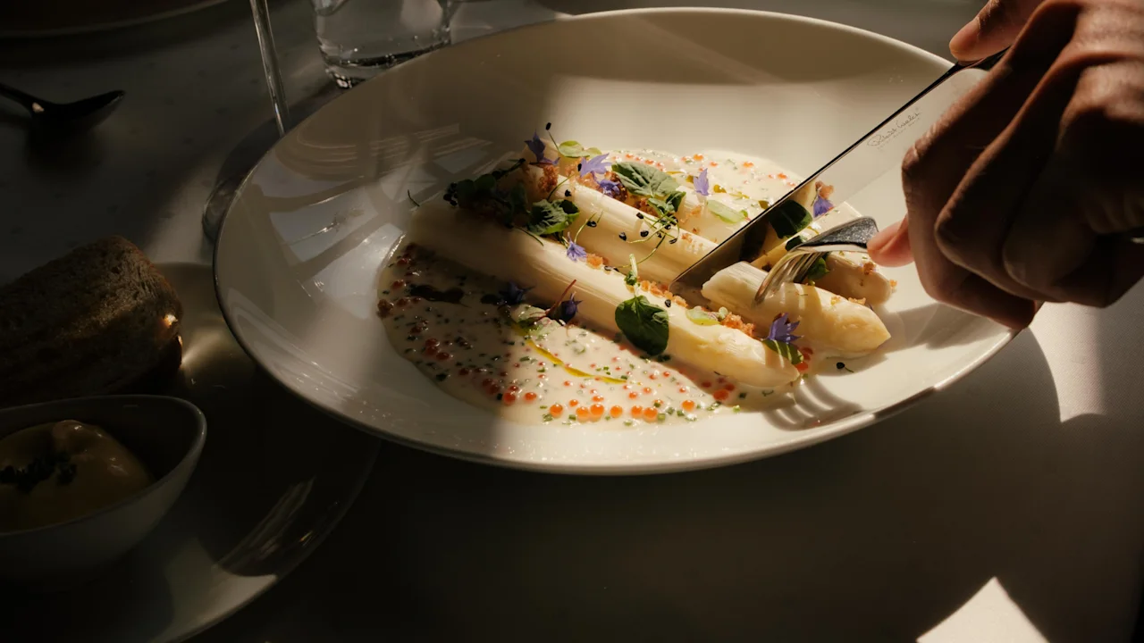 En person skär i en elegant maträtt med sparris, sås och ätbara blommor på en restaurang.