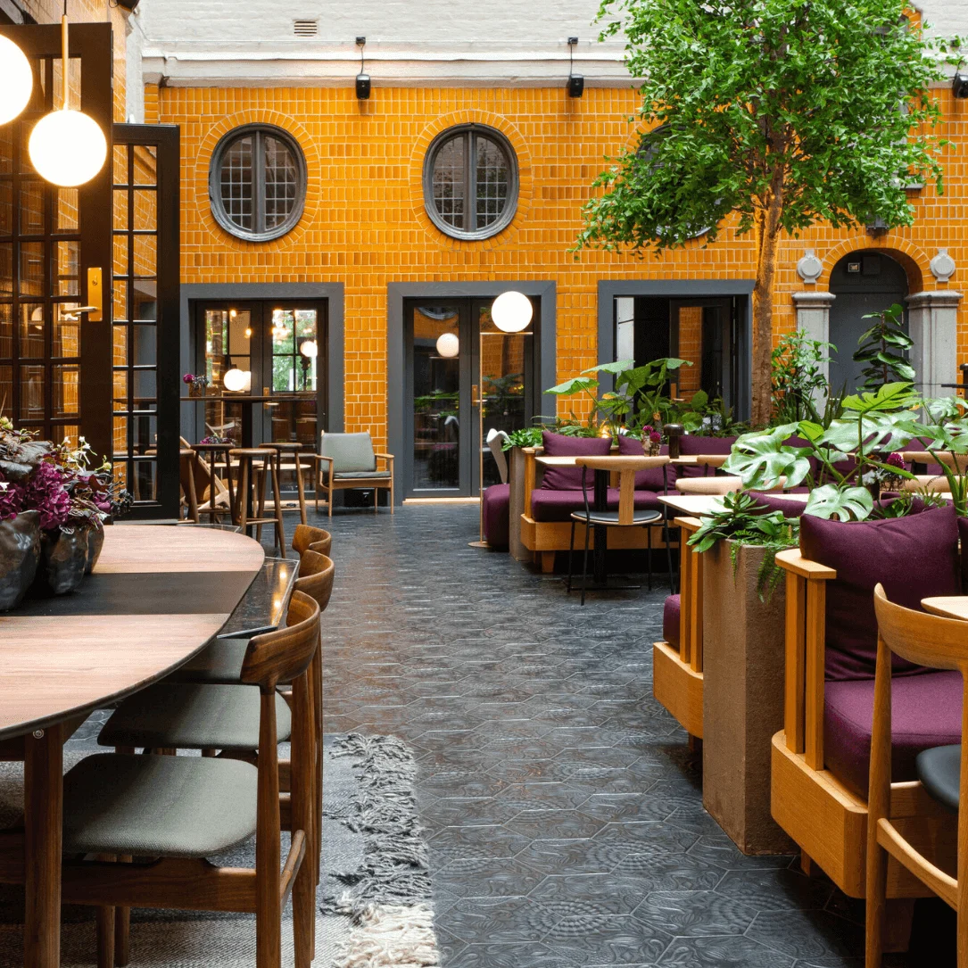 Tables and chairs are arranged in a modern, indoor café with orange-brick walls. Green plants decorate the space, while round lights hang from the ceiling, creating a cozy atmosphere.