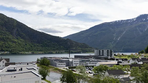Byggnader med utsikt över en lugn sjö, omgivna av frodiga gröna berg under en delvis molnig himmel. En smal flod rinner mellan strukturerna och förstärker den lugna atmosfären.
