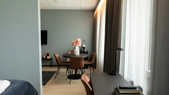 Et moderne hotellrom med en spiseplass med et rundt bord og brune stoler, samt en arbeidsplass med et skrivebord ved et vindu.