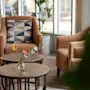 En mysig hotellounge med två fåtöljer, runda träbord och en vas med blommor, badad i naturligt ljus.