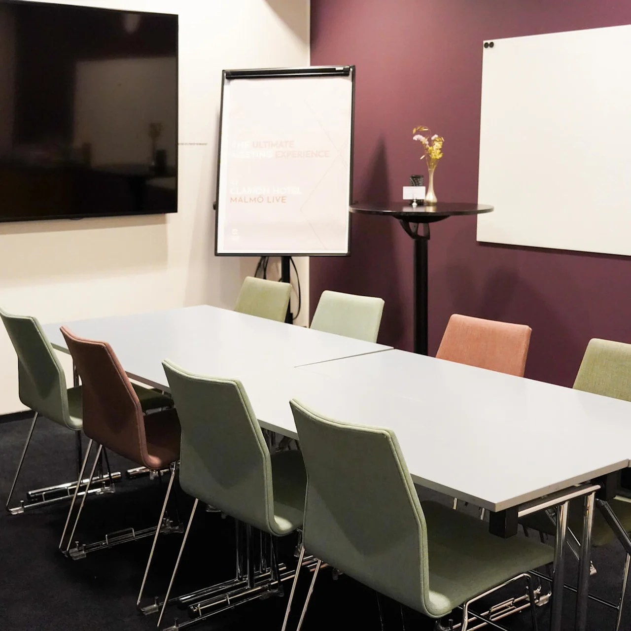 Et møterom med et bord omgitt av fargerike stoler. En flippover står med teksten: "The Ultimate Meeting Experience, Clarion Hotel, Malmo Live." I nærheten er en stor skjerm og en whiteboard.