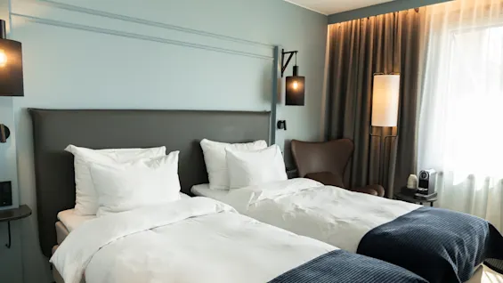 Et stilfullt hotellrom med to senger, skarpe hvite sengetøy, blå pledd og en lyseblå vegg. Myk belysning og en kaffemaskin.