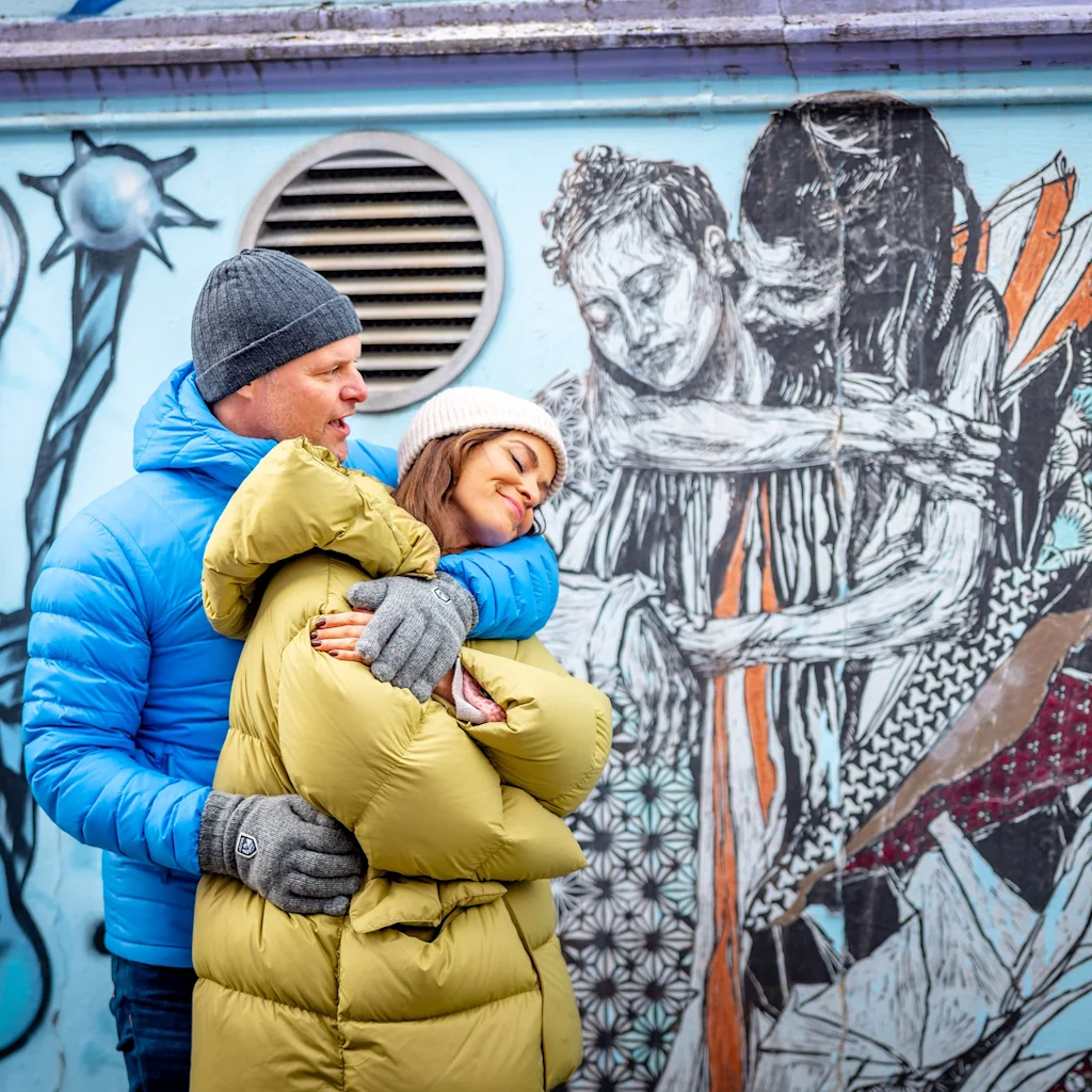 Kaksi ihmistä talvivaatteissa syleilee lämpimästi värikkään seinämaalauksen edessä, joka esittää syleilyä.