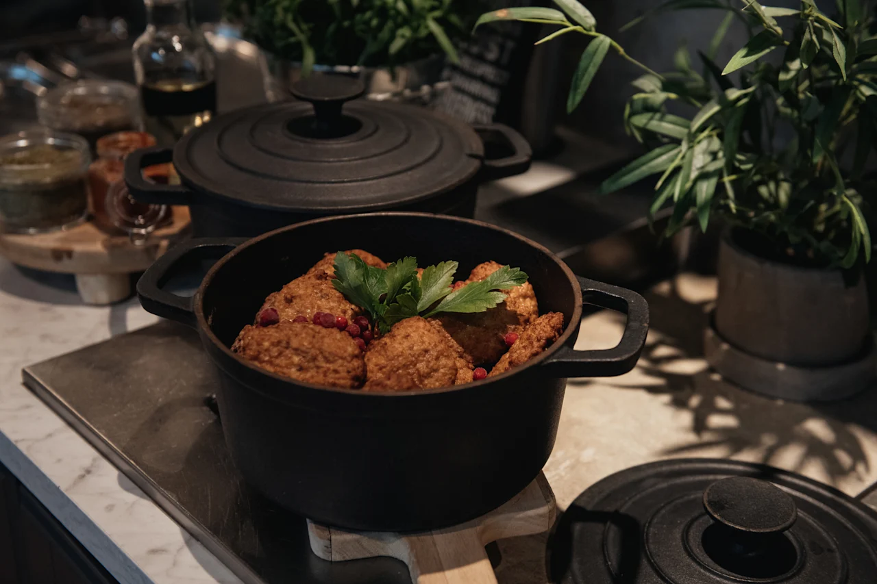 En sort gryde fyldt med tilberedt mad, pyntet med røde bær og grønne urter, på en disk med andre gryder og planter.