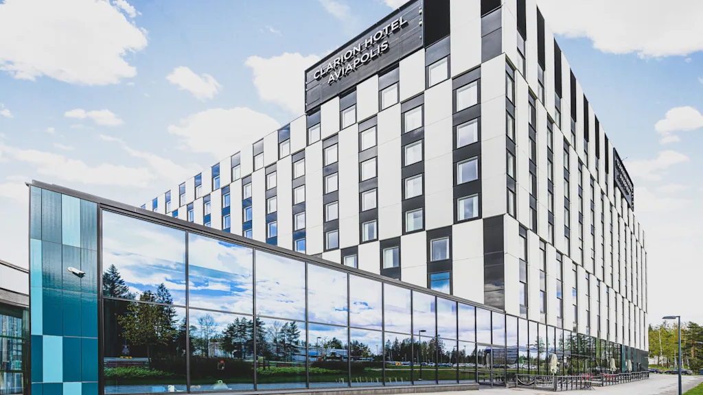 En flerplanshotellbyggnad med rutfasad står under blå himmel. Stora fönster reflekterar träd. Text: "CLARION HOTEL AVIAPOLIS."