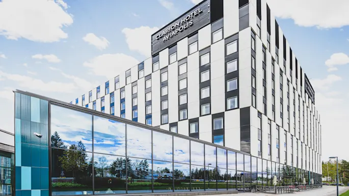 En fleretasjes hotellbygning med rutefatature står under en blå himmel. Store vinduer reflekterer trær. Tekst: "CLARION HOTEL AVIAPOLIS."