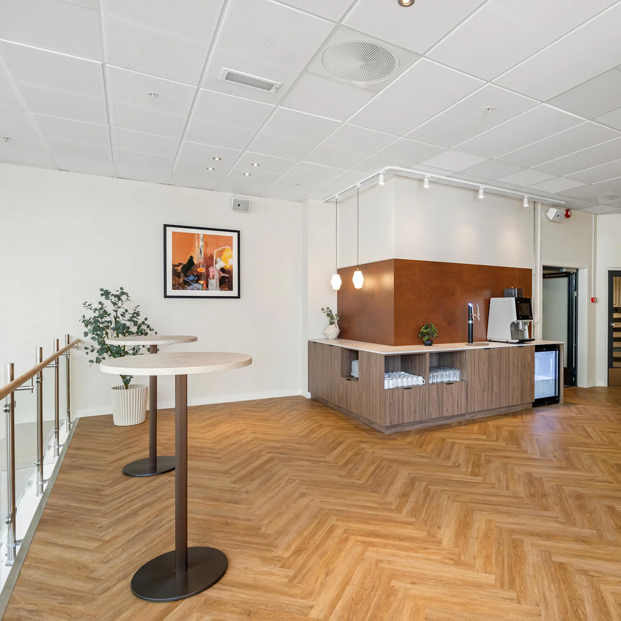A modern reception area with a wooden front desk, two high tables, and a framed picture on the wall, inside a well-lit, spacious room with herringbone flooring.