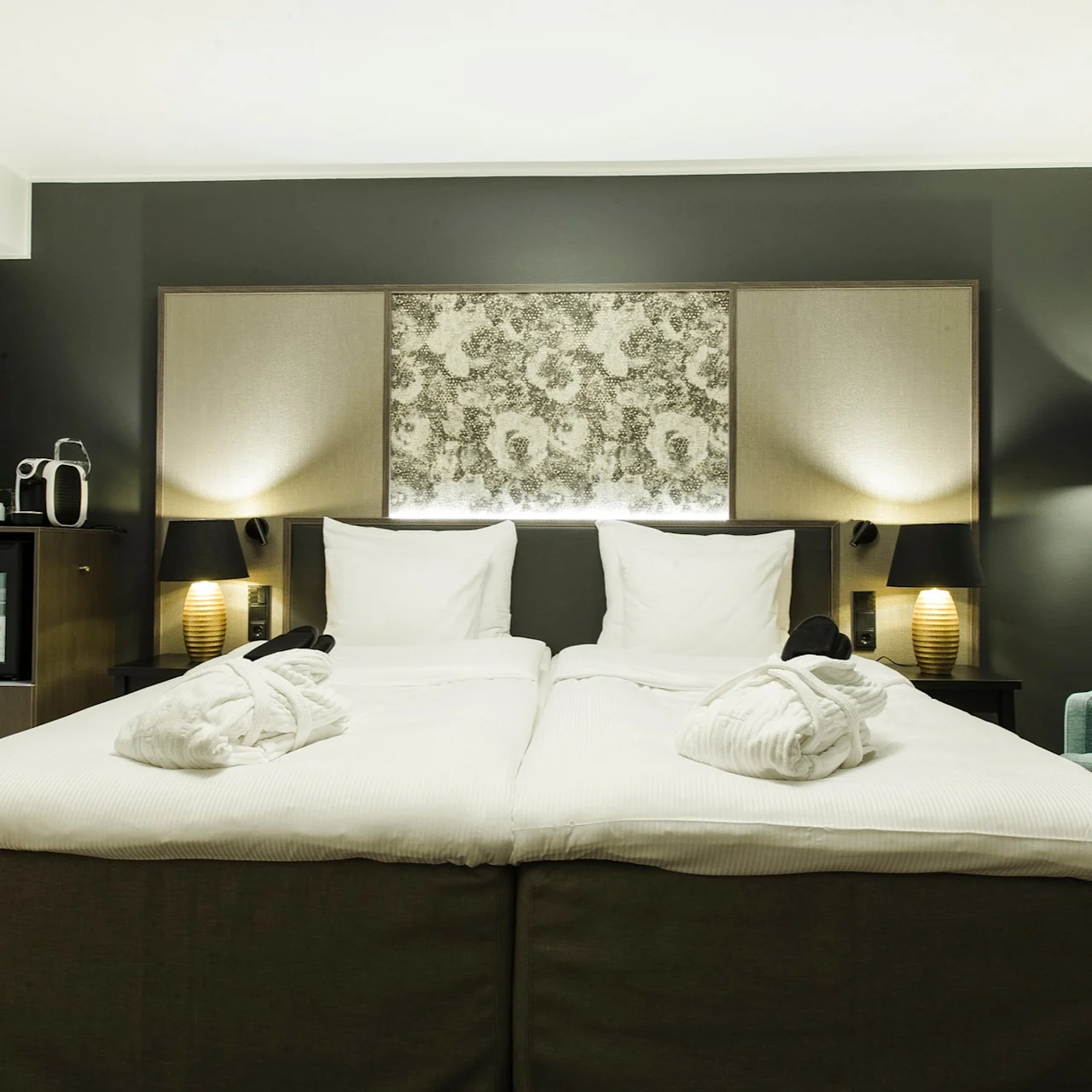 A neatly made double bed with white linens and two folded bathrobes, flanked by lamps, in a modern hotel room. A teal armchair and a minibar with a coffee machine are nearby.