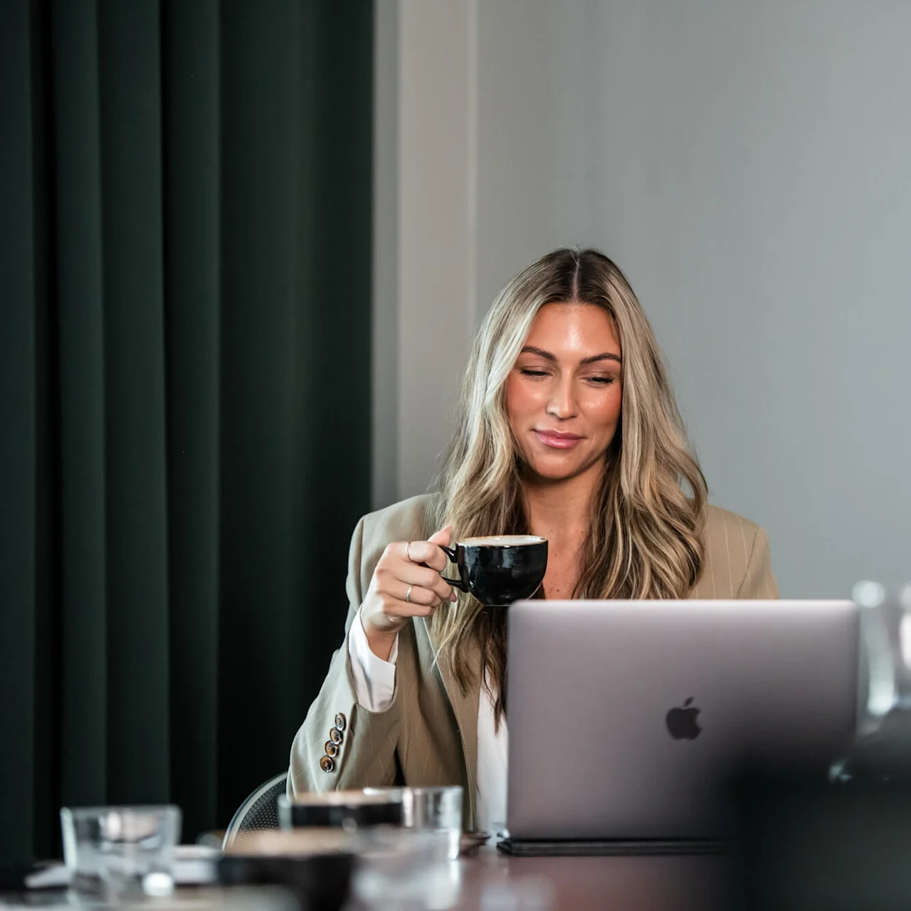 A woman, sitting at a table, sips from a black cup while using a laptop. The setting includes blurred glassware in the foreground and dark curtains.