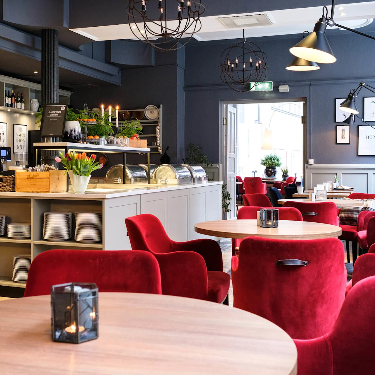 A cozy cafe interior with red velvet chairs and wooden tables, lit with warm lighting and adorned with plants.