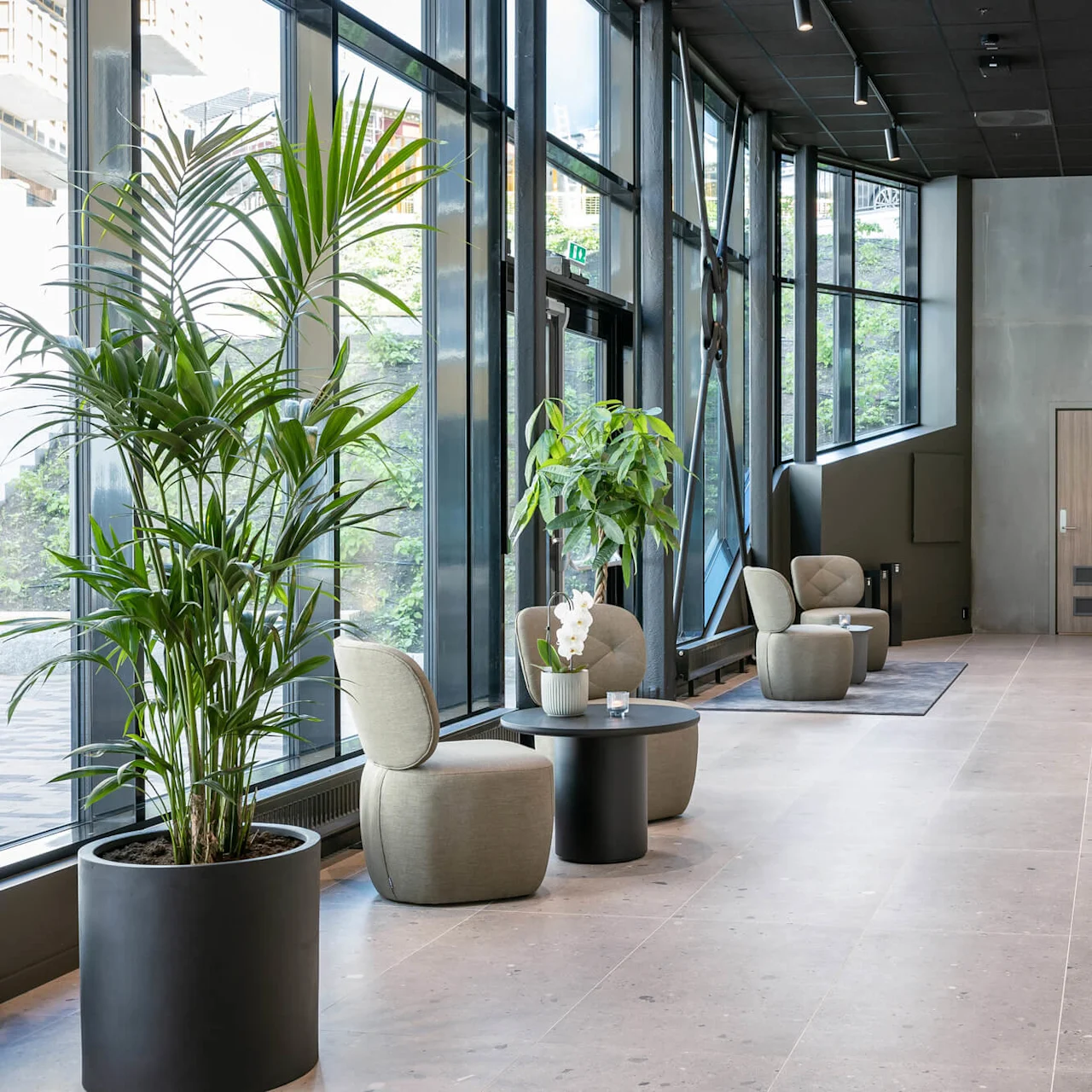Moderne lobby med grønne lænestole rundt om runde borde, ved siden af store vinduer og potteplanter. Glatte vægge og gulv skaber en minimalistisk atmosfære. Dør markeret "2b Kontor."