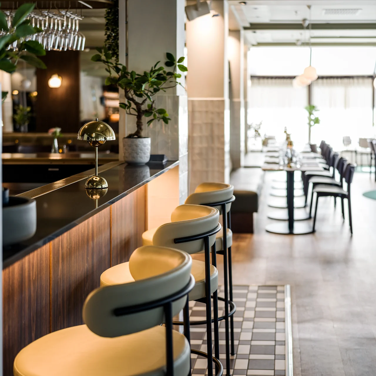 Elegant barstools line a sleek counter adorned with a gold lamp and plants, leading into a spacious, brightly-lit restaurant with tables and chairs, featuring large windows.