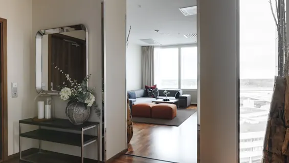 En modern hall med ett dekorativt konsolbord och spegel, som leder in till ett ljust vardagsrum med soffa och stora fönster.