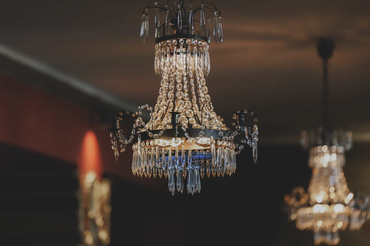 A crystal chandelier hangs from the ceiling, casting a warm glow in a dimly lit, elegant room, with another chandelier visible in the background.