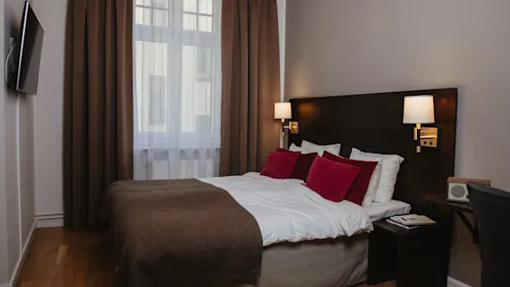 En pænt redt seng med hvidt og brunt sengetøj, suppleret med røde puder, står i et rent, moderne soveværelse. Den flankeres af sengeborde med lamper under dæmpet belysning.
