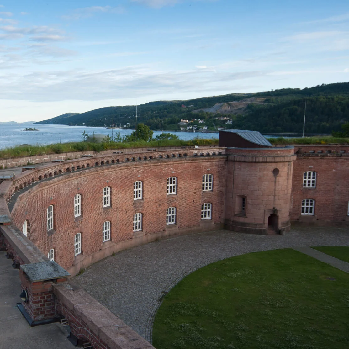 En buet murstensborg med utsikt over et rolig vann, omgitt av gresskledde åser og fjerne skoger under en blå himmel med løse skyer.