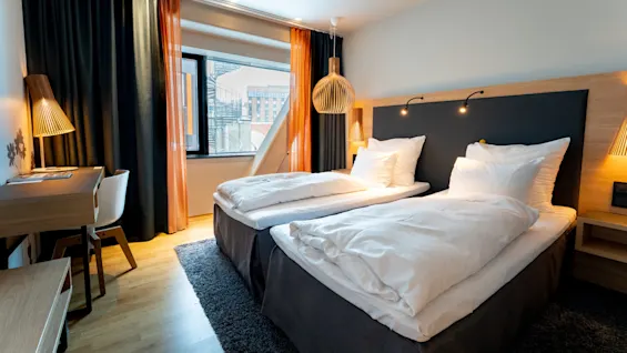 To pænt opredte enkeltsenge står side om side under væglamper, i et lyst, moderne rum med skrivebord og vindue med udsigt over byen, indrammet af gardiner.