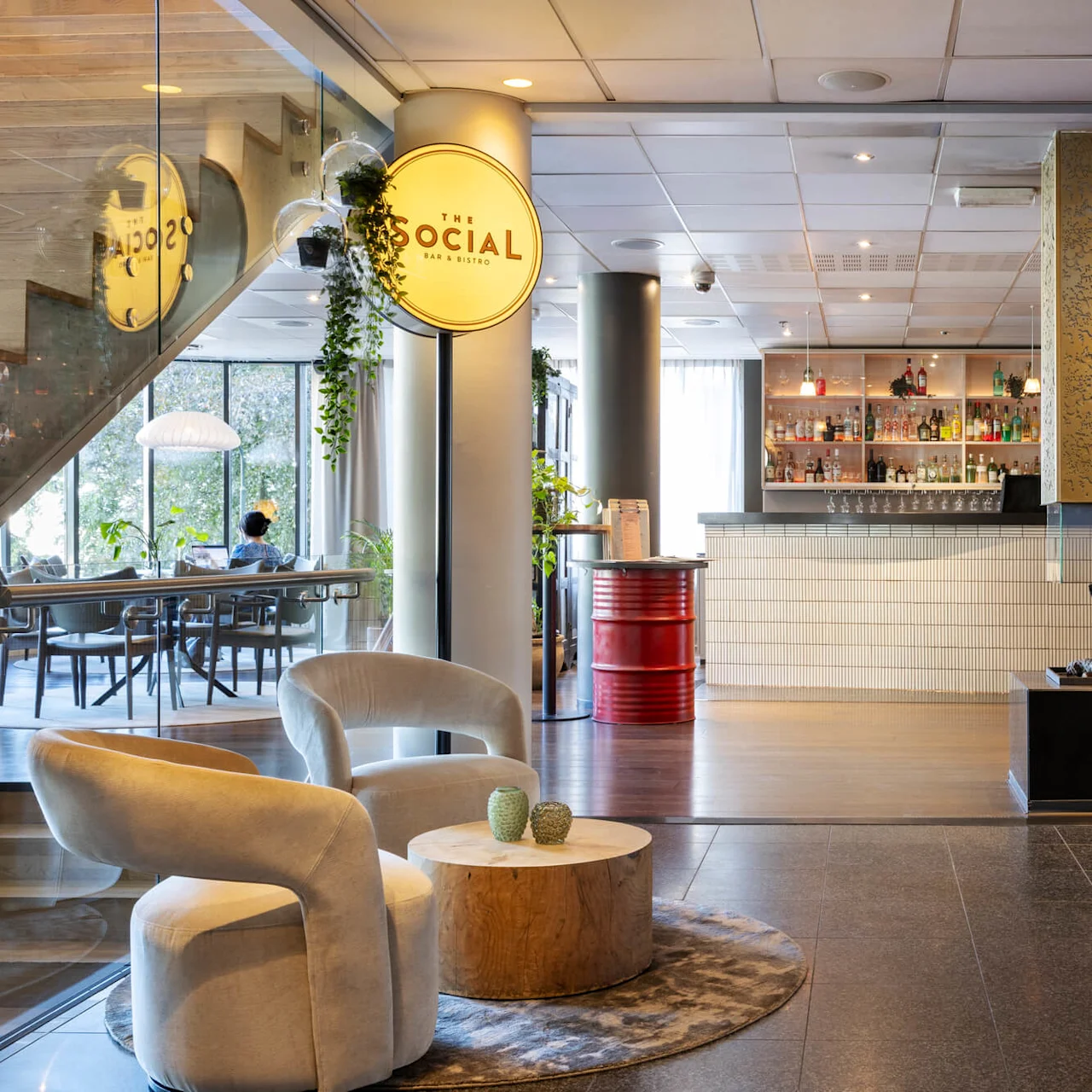 Two chairs and a round table sit in a modern lobby. Nearby, a sign reads "THE SOCIAL BAR & BISTRO" next to a bar and fireplace. Elevator doors visible.