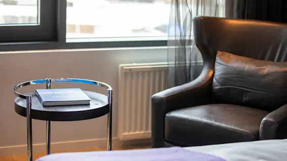 Et rundt bord har en bok med tittelen "HOTELLIELÄMÄ." En sort lenestol står i et solrikt rom med en radiator og et vindu.