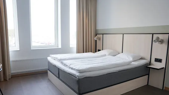 En pent ryddig dobbelseng med hvitt sengetøy står i et moderne rom, ved siden av store vinduer og beige gardiner, med tregulv og veggmonterte nattlamper.