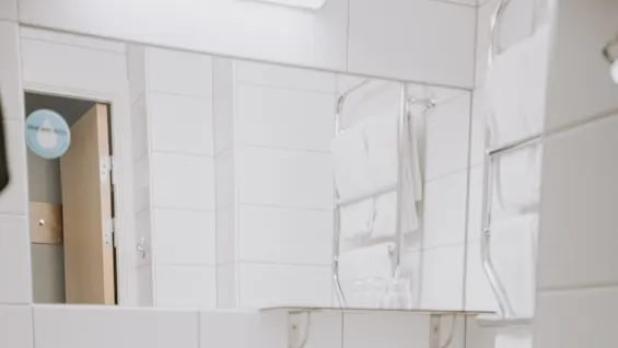 Et hvidt badeværelse har en vask med en sæbedispenser mærket "Hand Wash", et toilet, et vægspejl, håndklæder på et stativ og en metalaffaldsspand på et flisegulv.