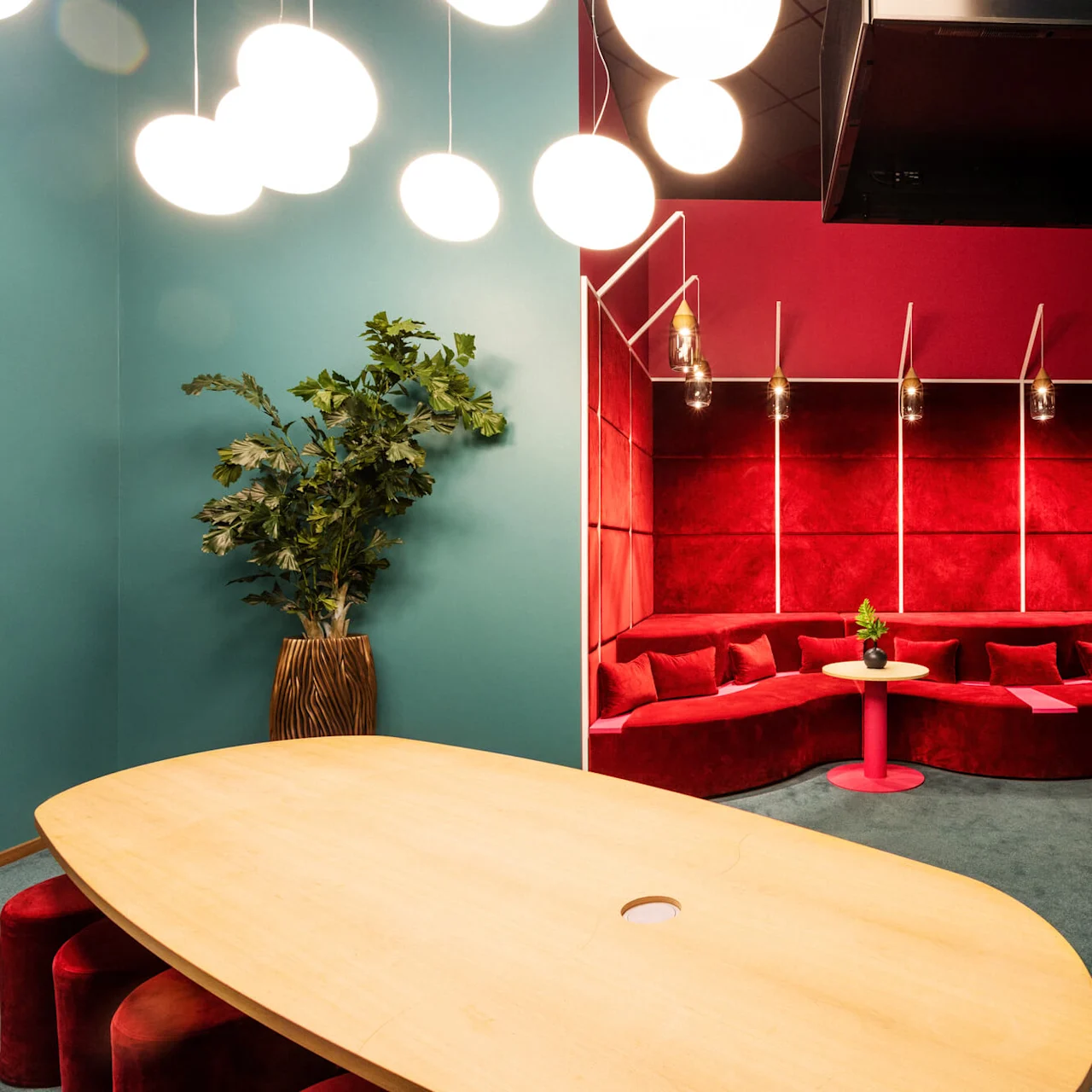 A wooden table is surrounded by red stools and lush plant decor, situated beside a vibrant red cushioned bench in a modern room with teal walls and circular lights.