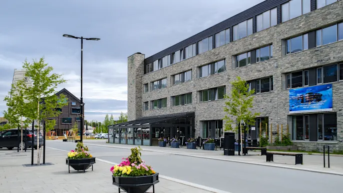 En modern stenbyggnad kantar en lugn gata, prydd med färgglada blomkrukor, kantad av unga träd och en stor banner. Molnig himmel och gatulampor ger stadskänsla.