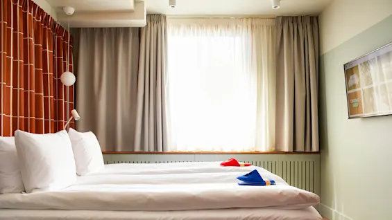 En seng med hvide lagner og to sæt hjemmesko står i et svagt oplyst rum med gardiner og et vægmonteret tv, som viser muligheder som "ROOM SERVICE" og "CHROMECAST".