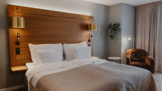 Et hyggeligt hotelværelse med en stor seng, sengegavl i træ og varm belysning, med en komfortabel lænestol og et strejf af grønt.