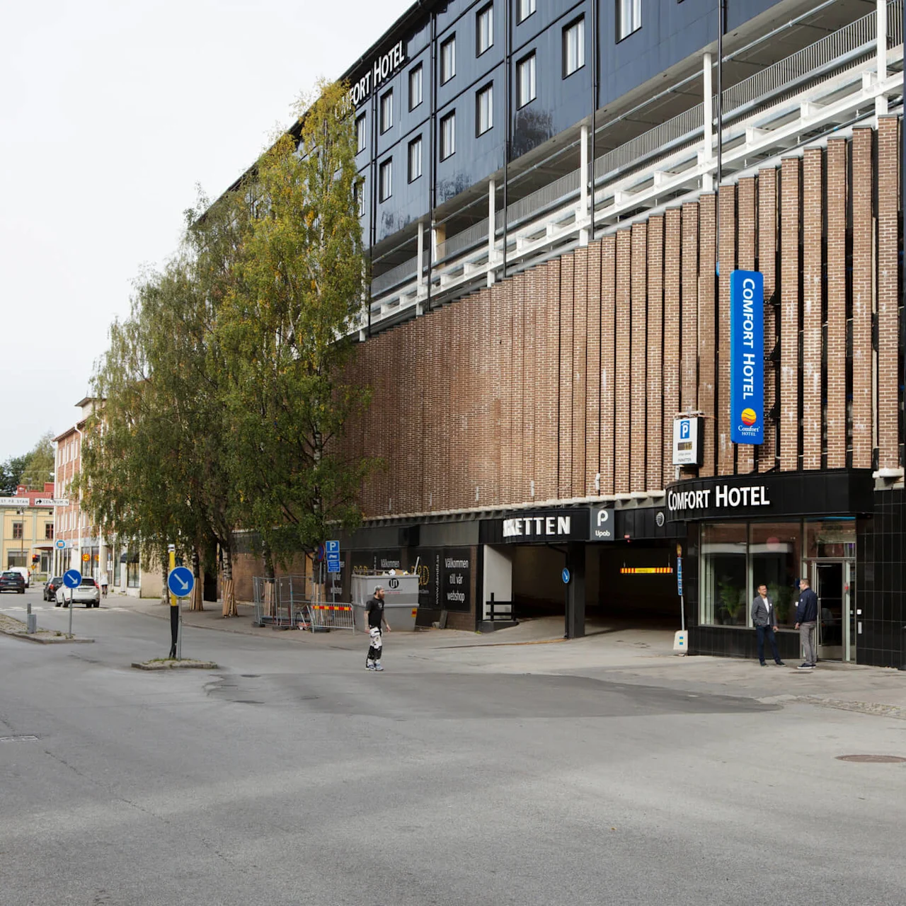 Comfort Hotel-byggnad med tegelvägg och blå skylt. Människor promenerar på gatan, med träd och parkeringsingång. Synlig text: "Comfort Hotel", "völkommen till vår webshöp".