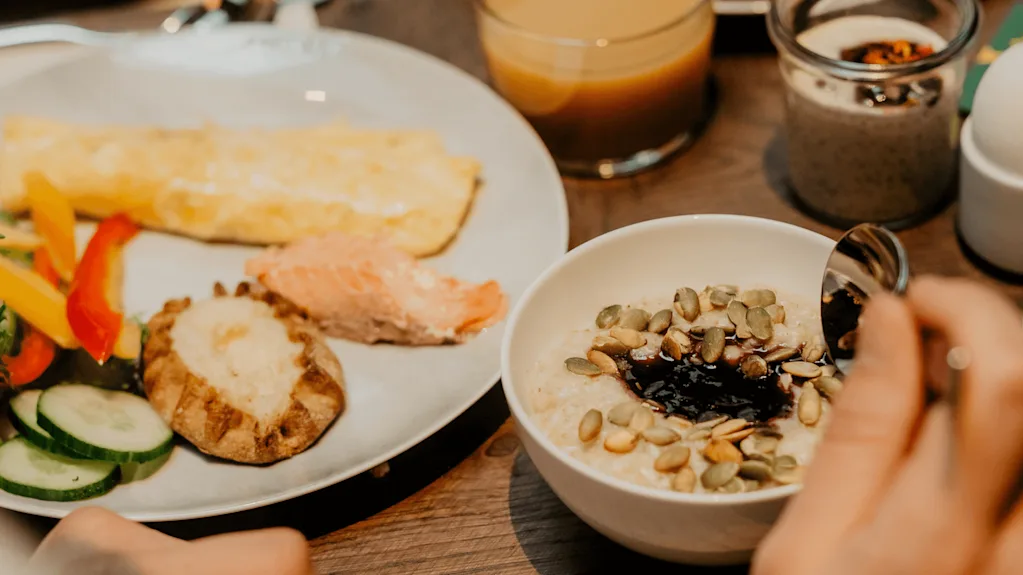 Bowl of porridge topped with seeds, surrounded by a hand holding a spoon. Nearby, a plate contains vegetables, salmon, omelette, and a potato. A glass of juice and yogurt are also visible.