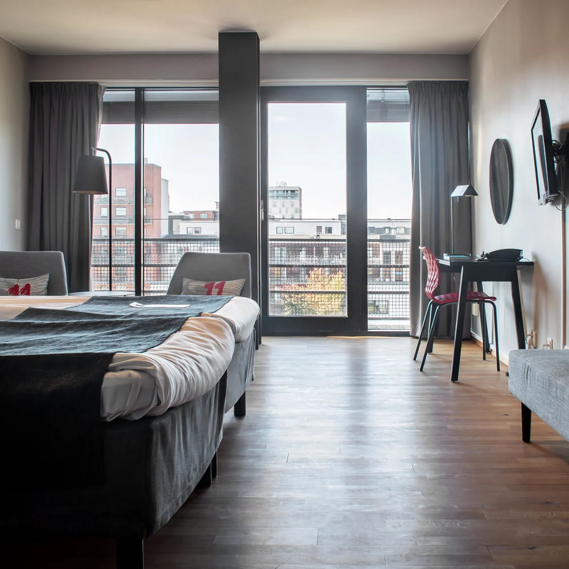 A hotel room with a neatly made bed, a desk, and a chair, overlooks a cityscape through a large window, featuring modern décor and minimalistic design.