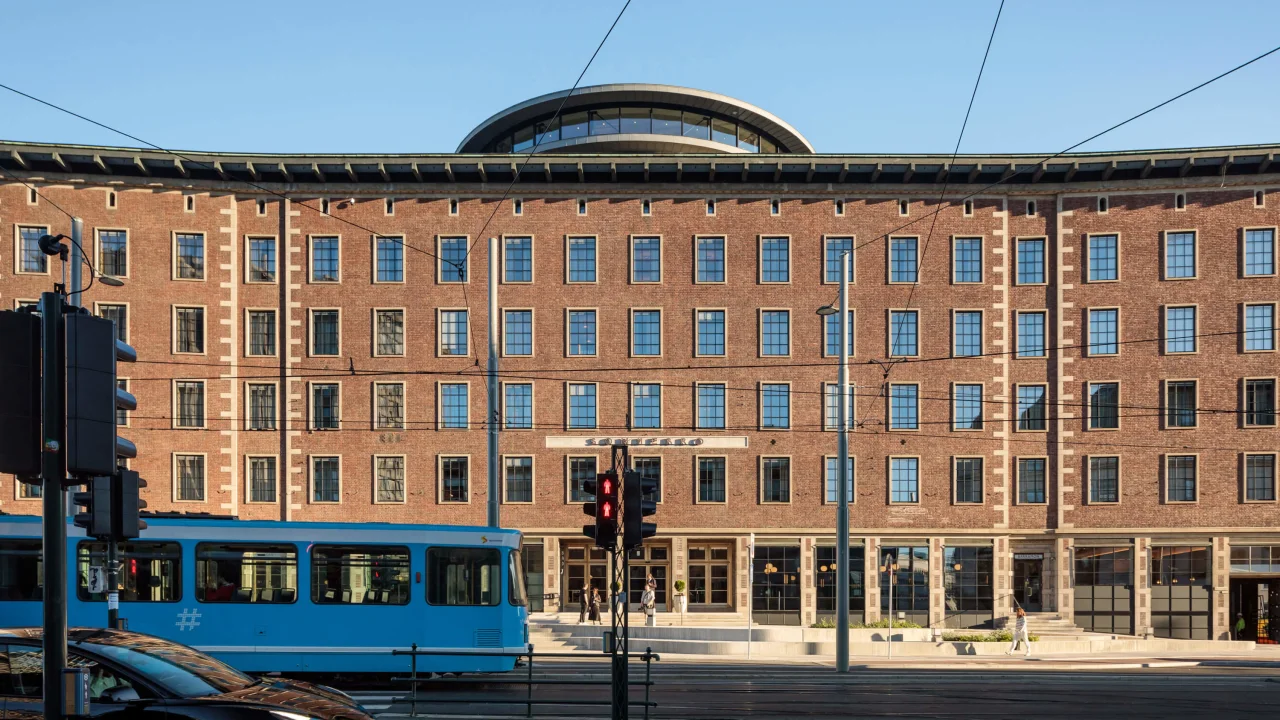 Mursteinsbygning med store vinduer bak en trikk på skinner; klar himmel.