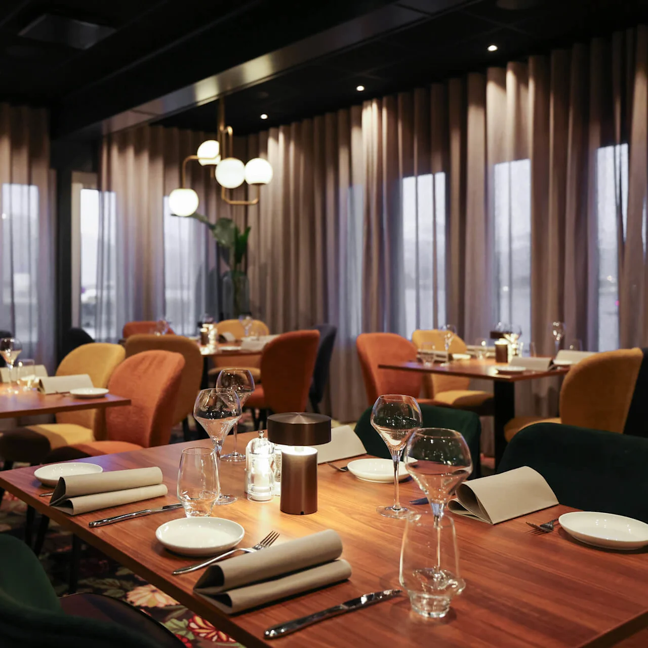 A restaurant interior with neatly set wooden tables, wine glasses, and folded napkins. Soft lighting and muted curtains create an intimate atmosphere.