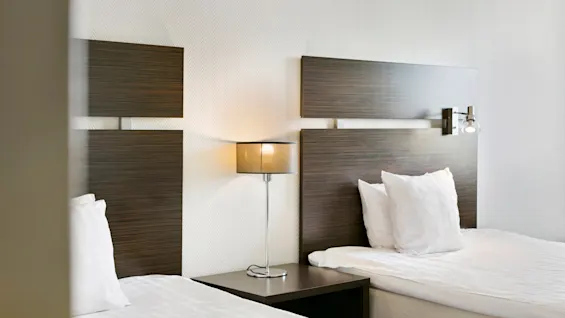 To pænt opredte senge med hvidt sengetøj står ved siden af et lille natbord med en moderne lampe, mod en væg med mørke trægavle i et hotelværelse.